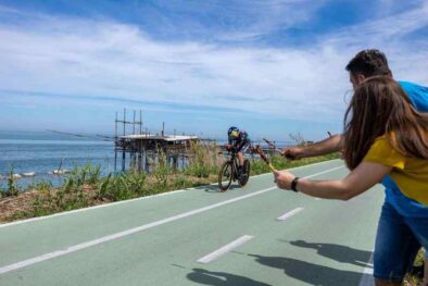 Un ciclista professionista in tenuta da cronometro pedala sulla Via Verde dei Trabocchi mentre due persone sorridenti a bordo pista gli offrono ironicamente degli arrosticini abruzzesi; sullo sfondo il mare Adriatico e un tipico trabocco in legno.