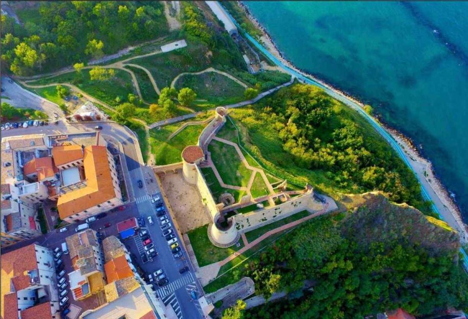 Una spettacolare veduta aerea perpendicolare del Castello Aragonese di Ortona. L'immagine mette in risalto la pianta trapezoidale della fortezza, circondata da prati verdi curati e sentieri geometrici. Sulla destra, la scogliera digrada verso il mare Adriatico dalle sfumature turchesi, mentre sulla sinistra si vedono i tetti del borgo e le auto parcheggiate lungo la strada costiera.
