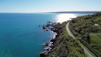 Una spettacolare veduta aerea della pista ciclabile "Via Verde" lungo la Costa dei Trabocchi a Ortona. Il percorso asfaltato si snoda sinuoso tra la fitta vegetazione collinare e la scogliera frastagliata a picco sull'Adriatico. In lontananza, un trabocco svetta tra le acque turchesi e cristalline, mentre il sole si riflette intensamente sul mare, creando una scia luminosa che accompagna il cammino dei ciclisti.