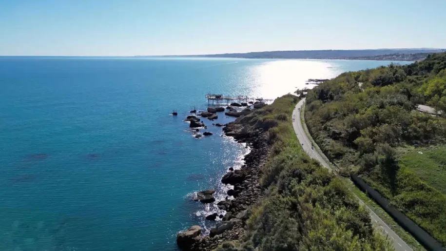 Una spettacolare veduta aerea della pista ciclabile "Via Verde" lungo la Costa dei Trabocchi a Ortona. Il percorso asfaltato si snoda sinuoso tra la fitta vegetazione collinare e la scogliera frastagliata a picco sull'Adriatico. In lontananza, un trabocco svetta tra le acque turchesi e cristalline, mentre il sole si riflette intensamente sul mare, creando una scia luminosa che accompagna il cammino dei ciclisti.