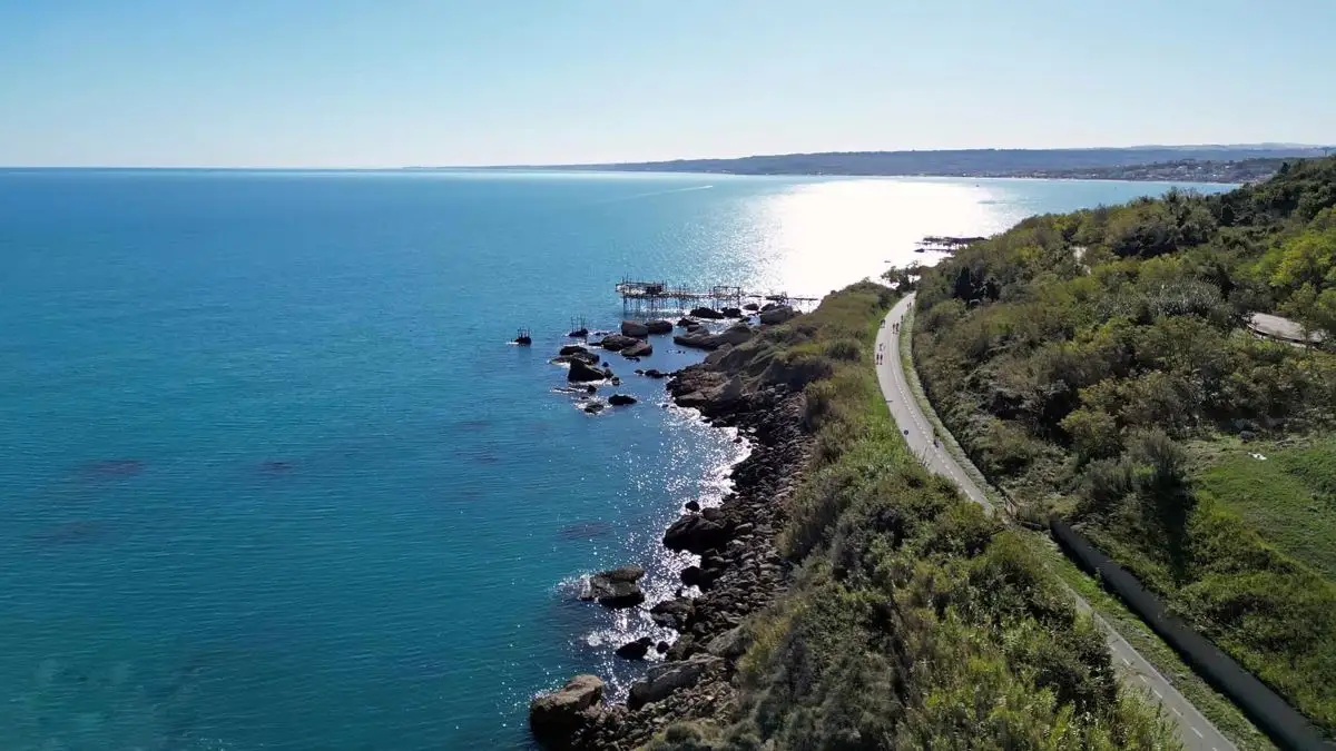 Una spettacolare veduta aerea della pista ciclabile "Via Verde" lungo la Costa dei Trabocchi a Ortona. Il percorso asfaltato si snoda sinuoso tra la fitta vegetazione collinare e la scogliera frastagliata a picco sull'Adriatico. In lontananza, un trabocco svetta tra le acque turchesi e cristalline, mentre il sole si riflette intensamente sul mare, creando una scia luminosa che accompagna il cammino dei ciclisti.