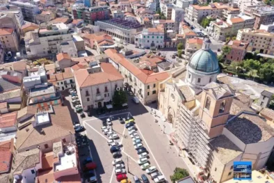 Una panoramica aerea zenitale di Piazza San Tommaso a Ortona, che mostra l'ampio parcheggio pubblico situato proprio di fronte alla Basilica di San Tommaso Apostolo. L'immagine evidenzia la disposizione ordinata dei posti auto e la vicinanza immediata alle strutture ricettive del centro storico, con i tetti in tegole rosse e i vicoli caratteristici che circondano l'area monumentale.
