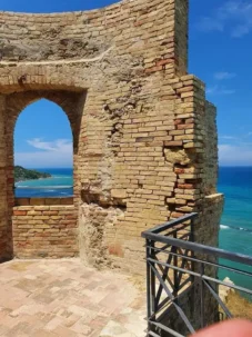 Veduta panoramica del mare Adriatico attraverso un'antica finestra ad arco in mattoni del Castello Aragonese. In primo piano, i dettagli materici della pietra storica e del pavimento in cotto, con una ringhiera in ferro che si affaccia sulla costa azzurra sotto un cielo limpido.