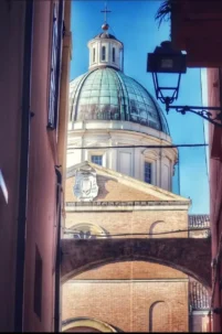 Veduta prospettica da un vicolo storico di Ortona che incornicia la grande cupola verde della Basilica di San Tommaso Apostolo, con un arco in mattoni in primo piano e un cielo azzurro sullo sfondo