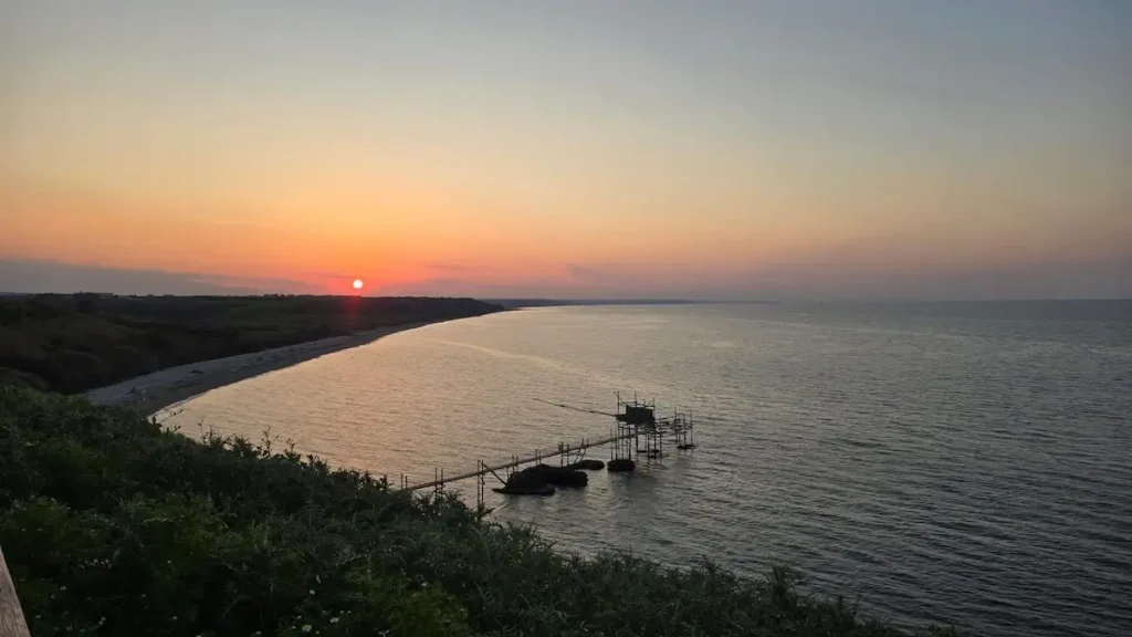 Veduta panoramica dall'alto della Riserva Naturale di Punta Aderci al tramonto, con un trabocco tradizionale proteso nel mare e la costa abruzzese all'orizzonte.