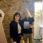 Foto ricordo di Lucia (a sinistra) e della scrittrice Giovanna Ceriotti (a destra, che regge il suo libro) posano insieme all'interno del B&B La Dimora di Tommaso, con lo sfondo di un'antica parete in pietra